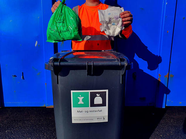Kundeveileder holder opp en grønn matavfallspose og en bleie. Foto.